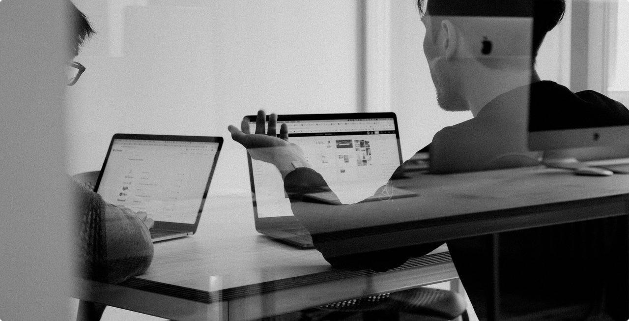 A black and white overhead shot shows two people sitting at a table, each using a tablet or laptop. Their faces are blurred, with the focus on the devices and their interaction.