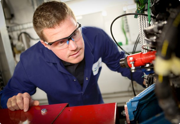 A mechanic wearing safety glasses and blue overalls leans over an engine, performing maintenance.