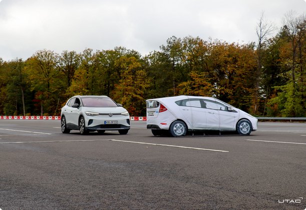 Two white test vehicles are driving on an asphalt test track with dashed markings and trees in the background.
