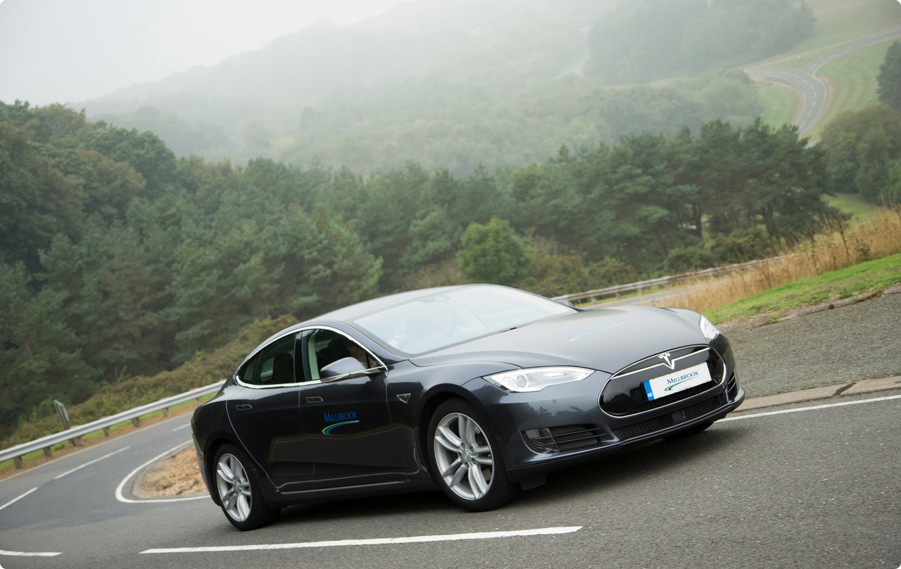 A dark, modern car drives on a winding road in a foggy, forested mountain landscape.