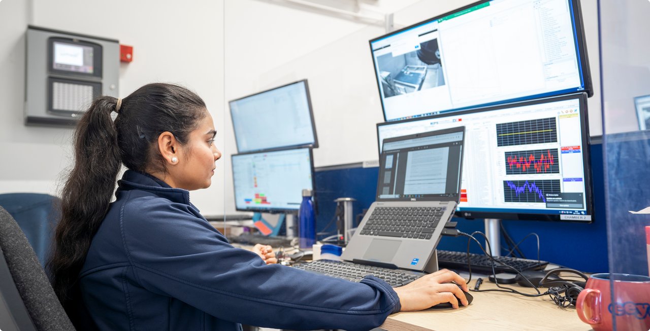 A woman is focused while working on a laptop in front of multiple screens displaying complex data and diagrams, in a modern office environment.