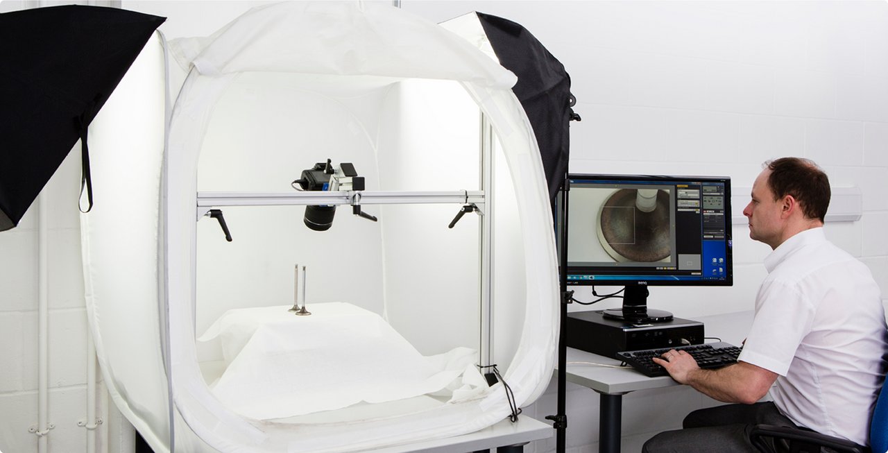 A man sits at a computer, looking at a monitor that displays a close-up of an object being photographed in a professional studio setup with lighting and backdrops.