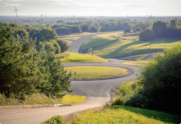 Eine erhöhte Ansicht einer kurvenreichen Test- oder Rennstrecke, die sich durch eine grüne, hügelige Landschaft schlängelt. Die Strecke ist umgeben von Bäumen und offenen Grasflächen, die im Sonnenlicht liegen. Im Horizont sind Umrisse einer Stadt oder Industrieanlage erkennbar.