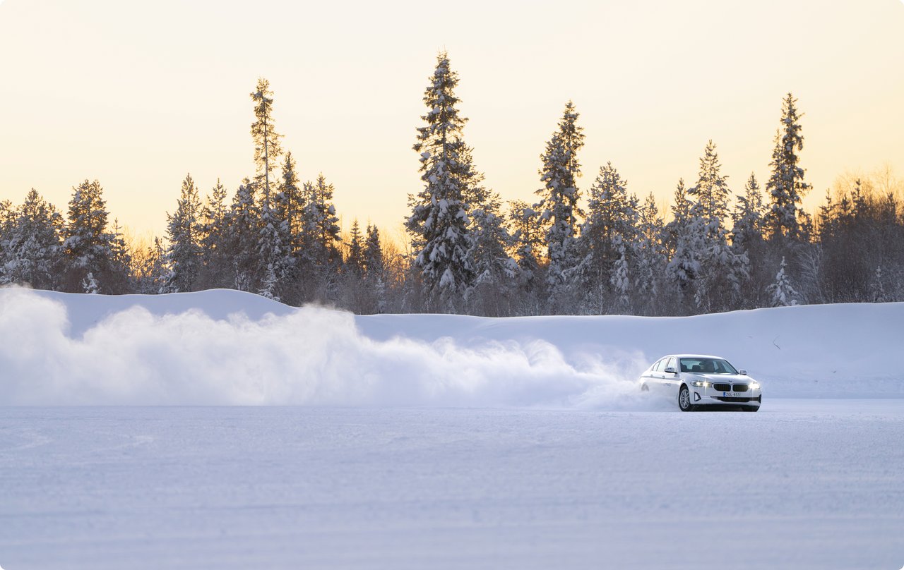 A white car is drifting on a vast snow-covered plain, kicking up a plume of snow, with a line of snow-dusted evergreen trees under a warm, low sun in the background.