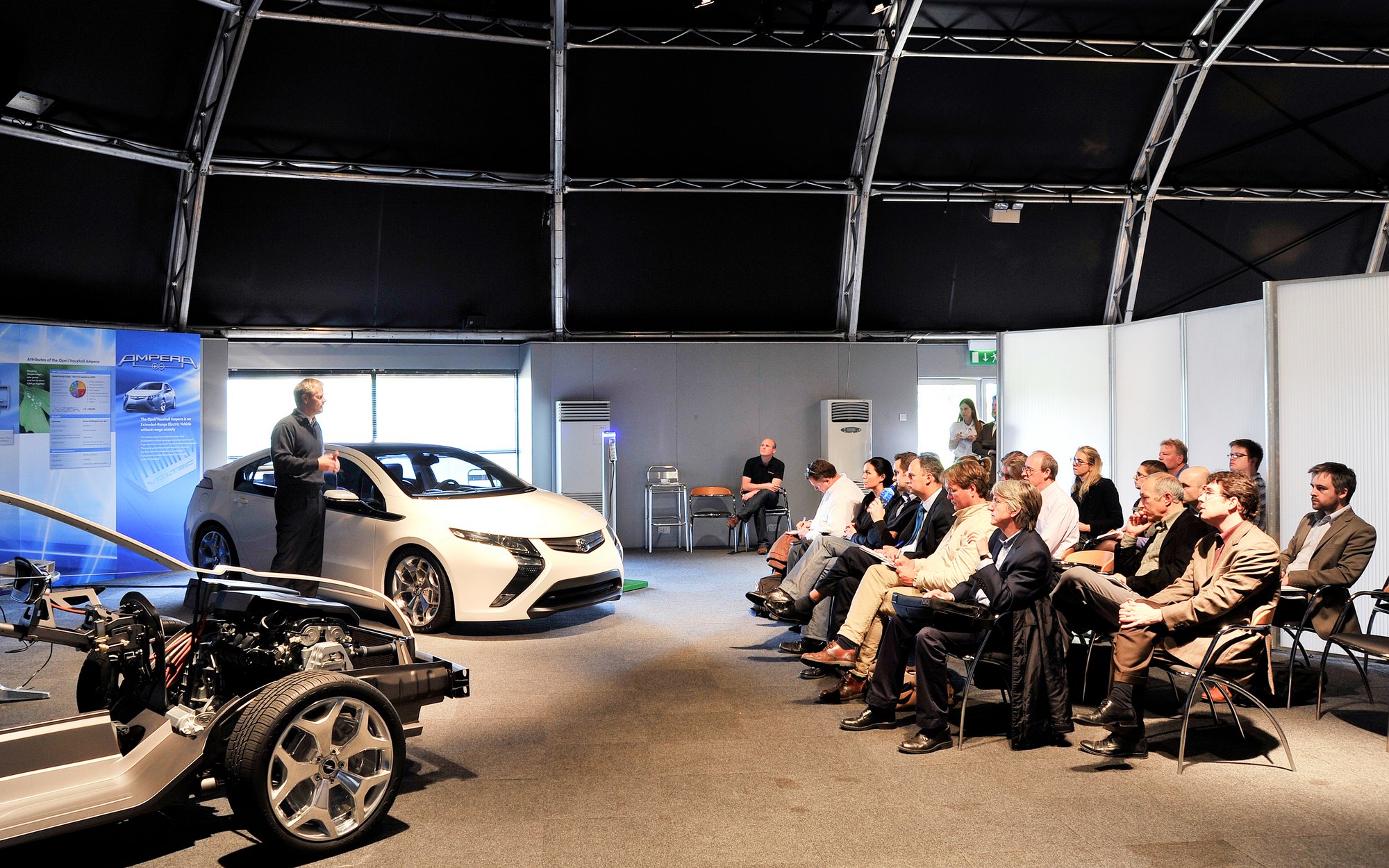 A group of people are seated or standing at a presentation in a large hall. A white car is displayed on a raised platform, with an exposed chassis visible in the foreground.