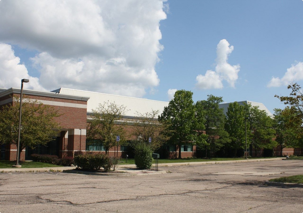An outdoor scene showing a gravel or unpaved area in the foreground with scattered trees and some plants in pots. In the background, there's a building with a reddish-brown facade and a large, modern white roof structure under a sky with both blue patches and white clouds.
