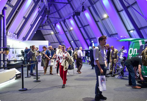 People are walking and mingling inside a large, modern exhibition or event hall with high, angular ceilings illuminated by purple and blue lighting. Various display stands and exhibits are visible in the background, suggesting a trade show or conference.