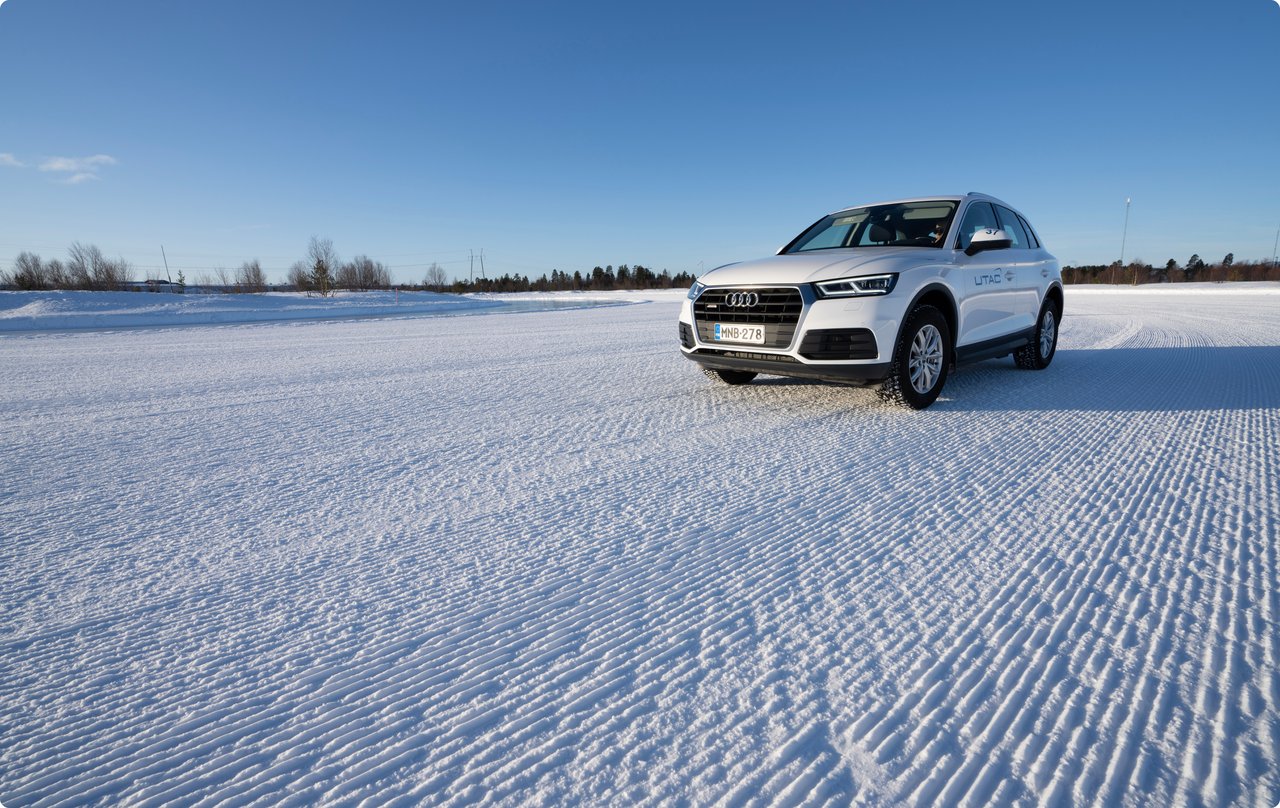 A white SUV is parked on a wide, flat, snow-covered surface under a clear blue sky. Distinct tracks or patterns are visible in the snow, suggesting test drives. Some indistinct buildings or trees are recognizable on the horizon.