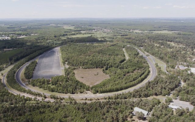 An aerial view of an expansive test facility featuring a large, U-shaped test track surrounded by dense forest. In the center of the U-shape is an open area, and a body of water is visible at the upper left edge of the curve.