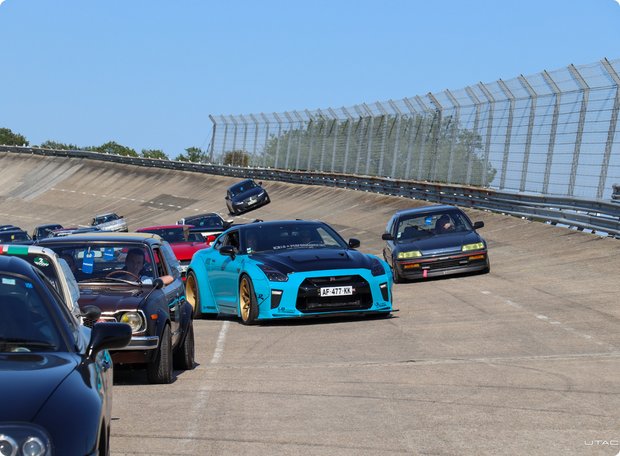 A turquoise sports car (Nissan GT-R) and other modified cars are parked on a gravel lot at an outdoor event, with a large wall or grandstand in the background.