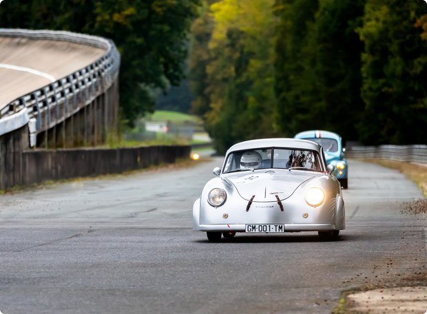 A historic white race car drives on a race track featuring a distinctive banked curve and a tree-lined straight. A second car is visible in the background. The track appears classic and close to nature.