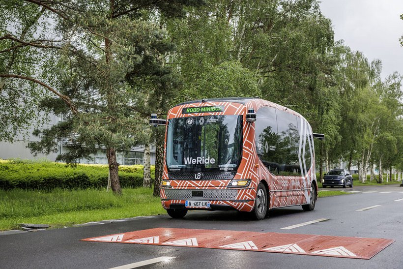 A modern, camouflage-patterned test bus is parked on a wet road, with a red mark visible on the asphalt in front of it. Trees line the road in the background.
