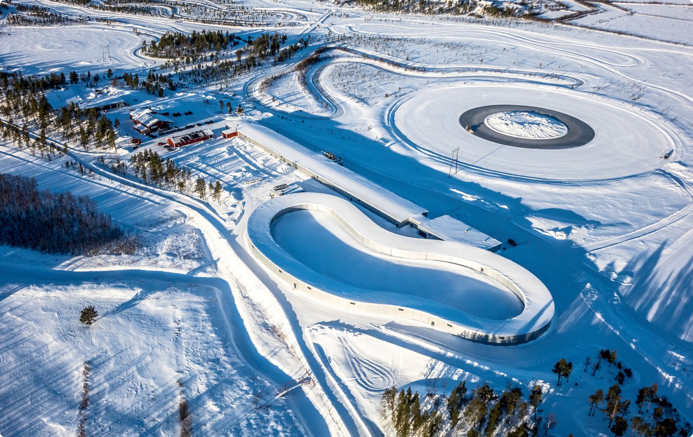 A winter aerial view shows a snow-covered test track or training area with multiple oval and circular paths laid out on an icy or snowy surface.