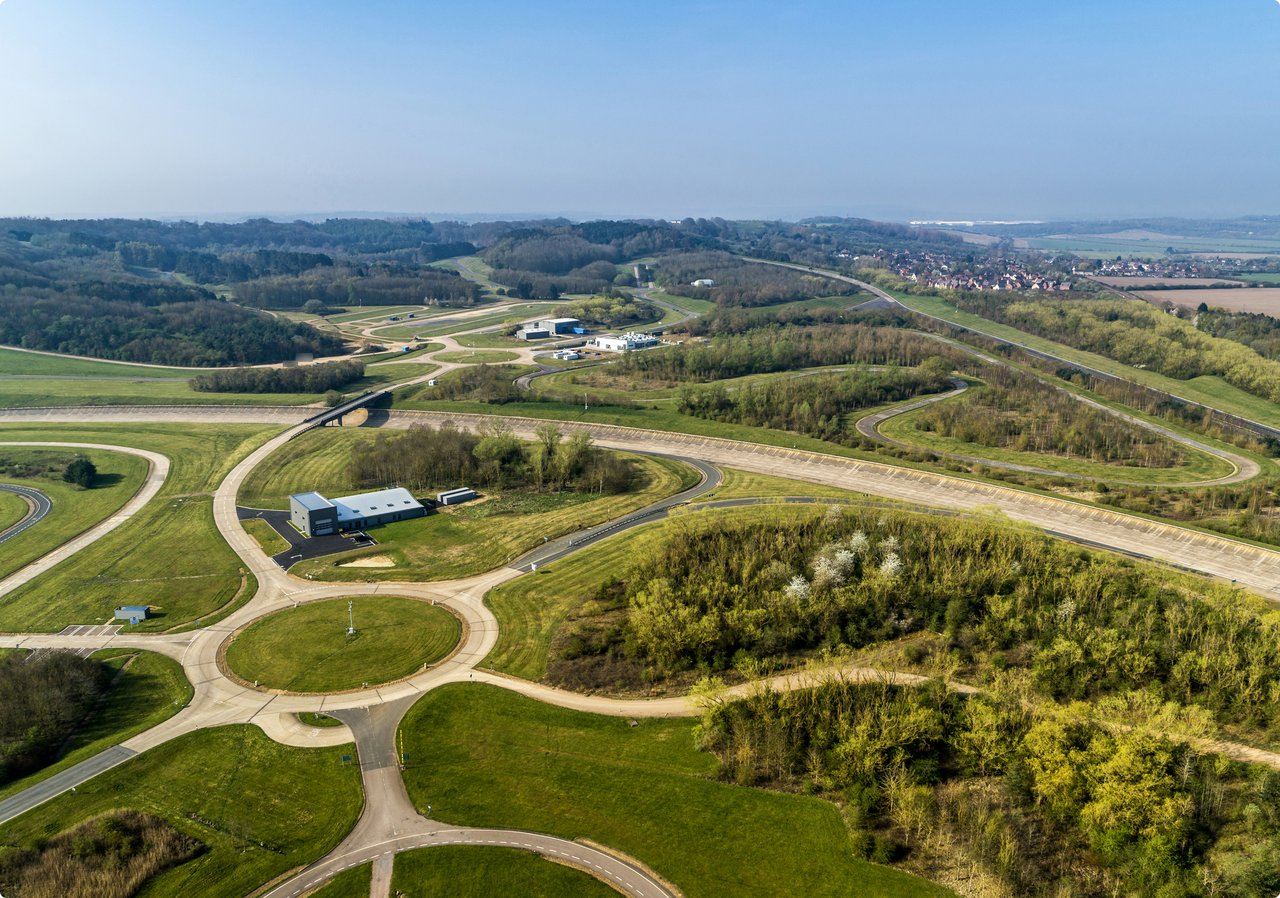 A wide aerial view shows a green landscape with a complex system of roads, roundabouts, and wooded areas under a clear blue sky.