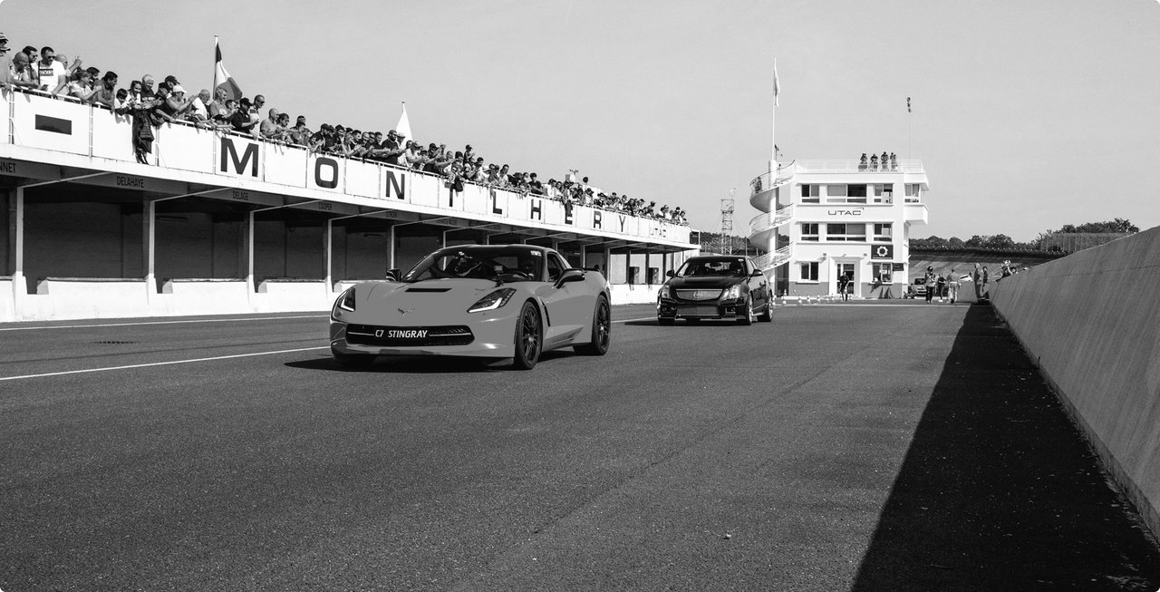 Black and white shot of a racetrack with an open-top sports car following another vehicle. Grandstands with spectators are on the left, and a tower is in the background.