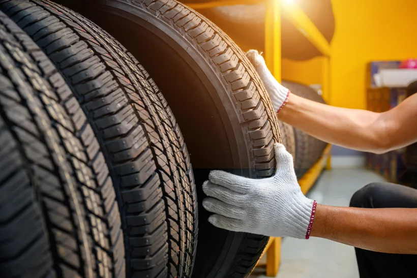 Close-up of a person's gloved hands taking a car tire from a shelf in a warehouse or workshop. In the background, more tires are visible on yellow shelves, and a warm ray of light falls into the room from above.