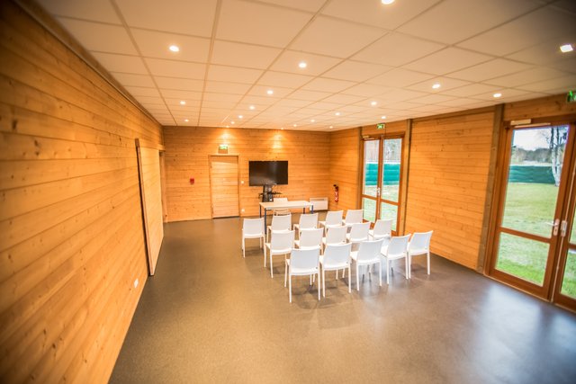 An empty modern meeting or conference room with light wood paneled walls, a dark grey floor, and a set of white chairs arranged in rows facing a screen and a small table at the front. Large windows on the right offer a view of a green outdoor area.