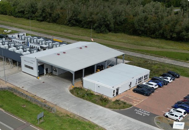 An aerial view shows a modern industrial facility with several buildings, a parking lot full of cars, and surrounding green areas.