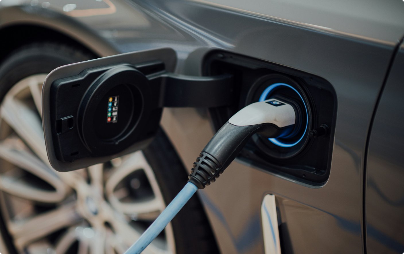 A close-up shot of an electric car being charged, showing the charging cable plugged into the car's port, which is illuminated with a blue light. The sleek design of the car and the charging connection are in sharp focus.