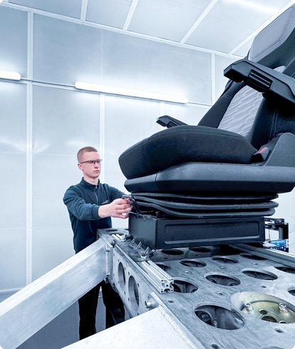 A technician with glasses and a dark shirt is adjusting an ergonomic driver's seat or a seat test bench in a clean, brightly lit laboratory or test room.