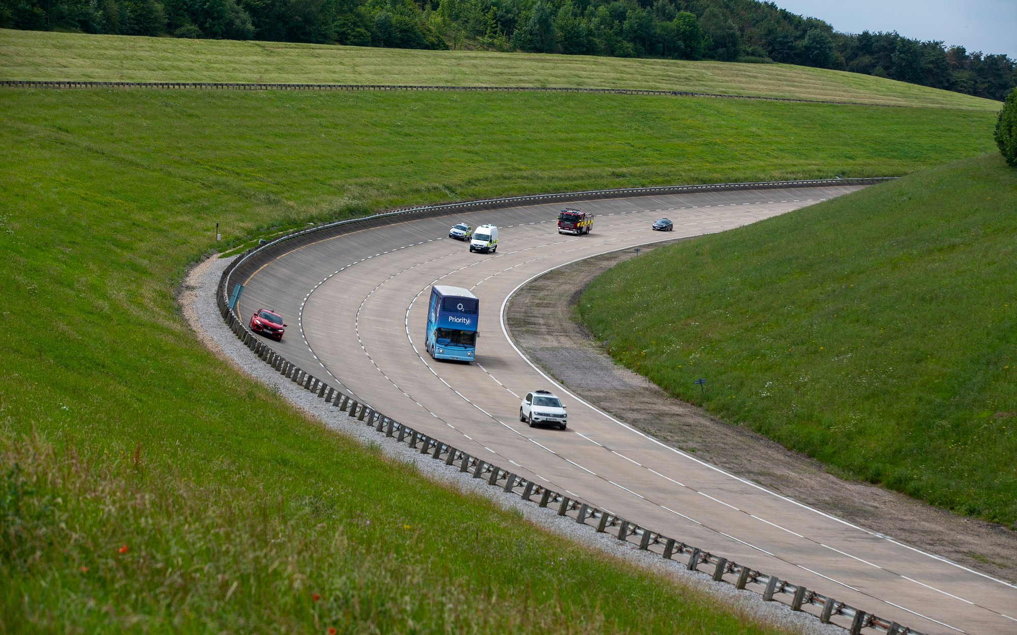 A group of cars, including a blue truck, drives on a winding test track through green hills and grasslands.
