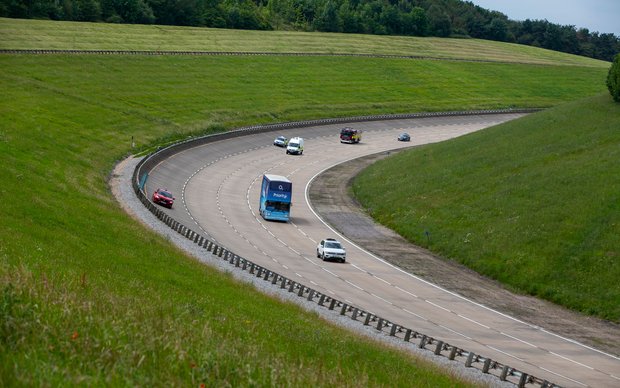 A group of cars, including a blue truck, drives on a winding test track through green hills and grasslands.