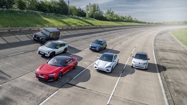 A collection of five diverse vehicles, including a red sports car, several SUVs, and a compact car, are parked or driving on a textured, unpaved test track with green grassy banks and trees in the background under a bright sky.
