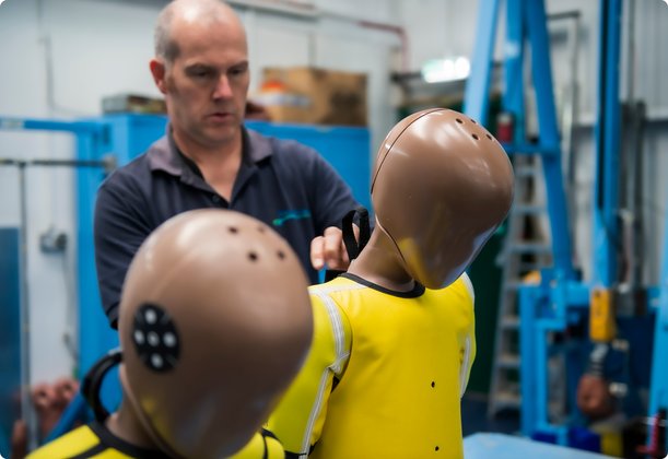 A bald and bearded technician in a dark polo shirt is preparing a crash test dummy. The dummy is brown and wears a yellow torso. Another dummy is blurred in the foreground.