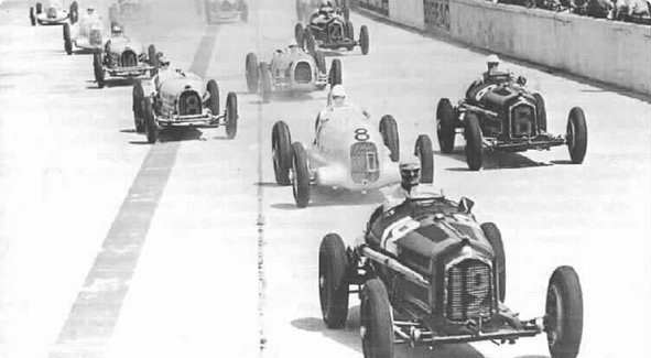 Une image historique en noir et blanc d'une rangée de voitures de course classiques sur une ligne de départ, prêtes pour une course.
