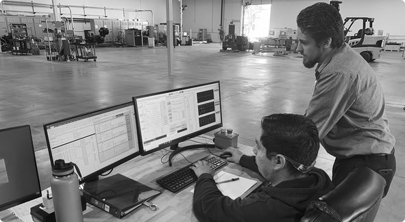 Deux personnes observent un poste de travail informatique dans un grand entrepôt industriel ou une usine avec des machines en arrière-plan.