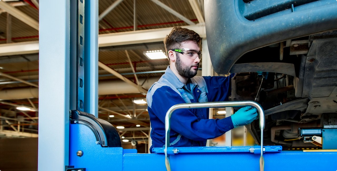A male technician wearing safety glasses and blue work overalls is working on the underside of a vehicle that is elevated on a blue car lift in a workshop.