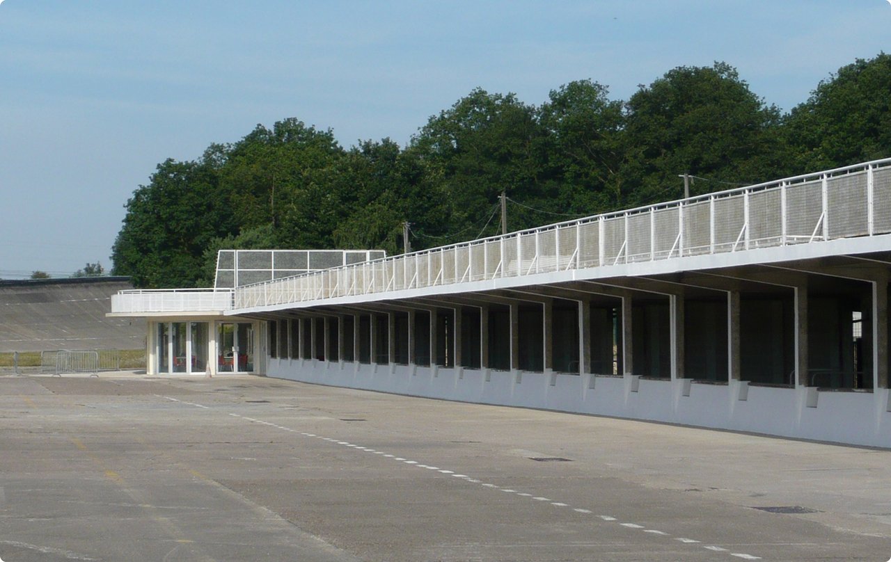 A long, white pit lane or garage building with an upper terrace at a race track. The asphalt in front of the building is empty, with trees and a blue sky in the background.
