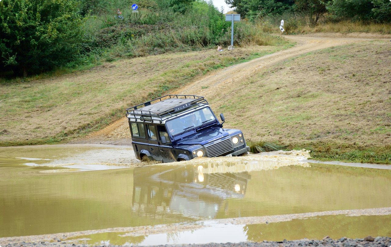 A dark off-road vehicle is partially stuck in a large, muddy puddle on rough terrain.
