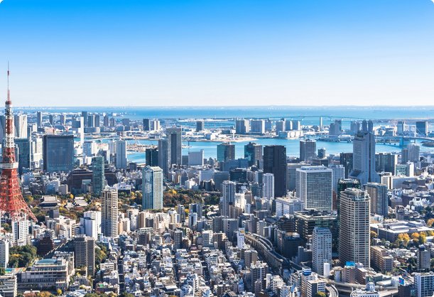 Vue aérienne étendue d'un horizon urbain densément peuplé, avec une proéminente tour de transmission rouge et blanche au premier plan à gauche, de nombreux immeubles de grande hauteur et une grande étendue d'eau avec un littoral lointain sous un ciel bleu clair.