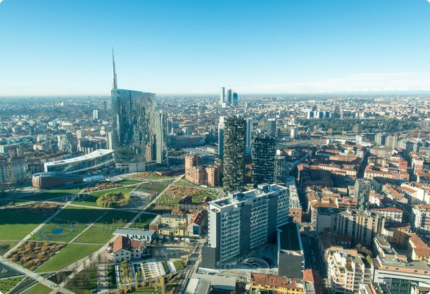Vue aérienne d'un paysage urbain moderne sous un ciel bleu clair, présentant de nombreux gratte-ciel et immeubles de grande hauteur, avec un grand espace vert en premier plan. La ville s'étend vers l'horizon avec un mélange d'architecture contemporaine et traditionnelle.