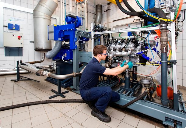 A technician in blue workwear kneels in front of complex industrial machinery consisting of pipes, valves, and gauges. He is wearing gloves and appears to be performing maintenance or inspection work. The environment is a workshop or laboratory.