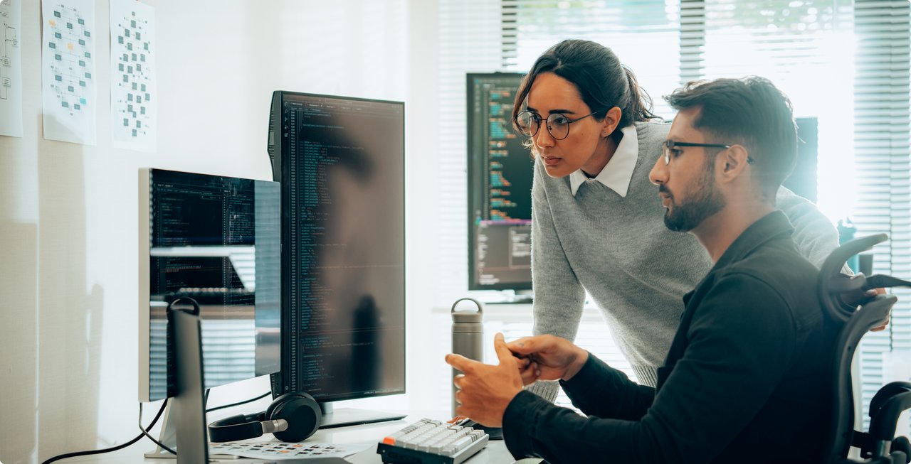 Two developers or IT specialists collaborate in front of multiple computer screens in a modern office; a woman leans over a man sitting at the desk as they jointly review code or data.