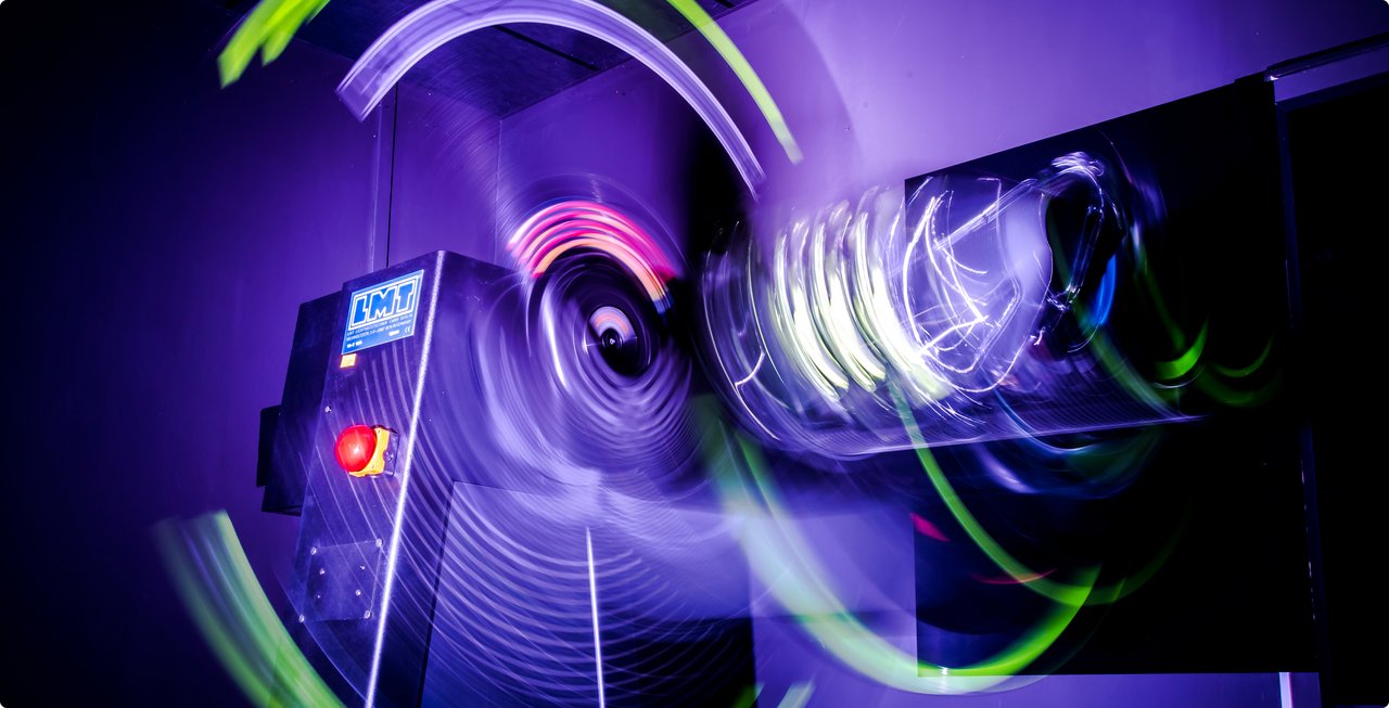 An abstract and vibrant image featuring bright purple and green light trails and reflections on a dark, possibly metallic, surface, creating a dynamic and futuristic effect. It might depict light art, a digital rendering, or a close-up of illuminated technology.