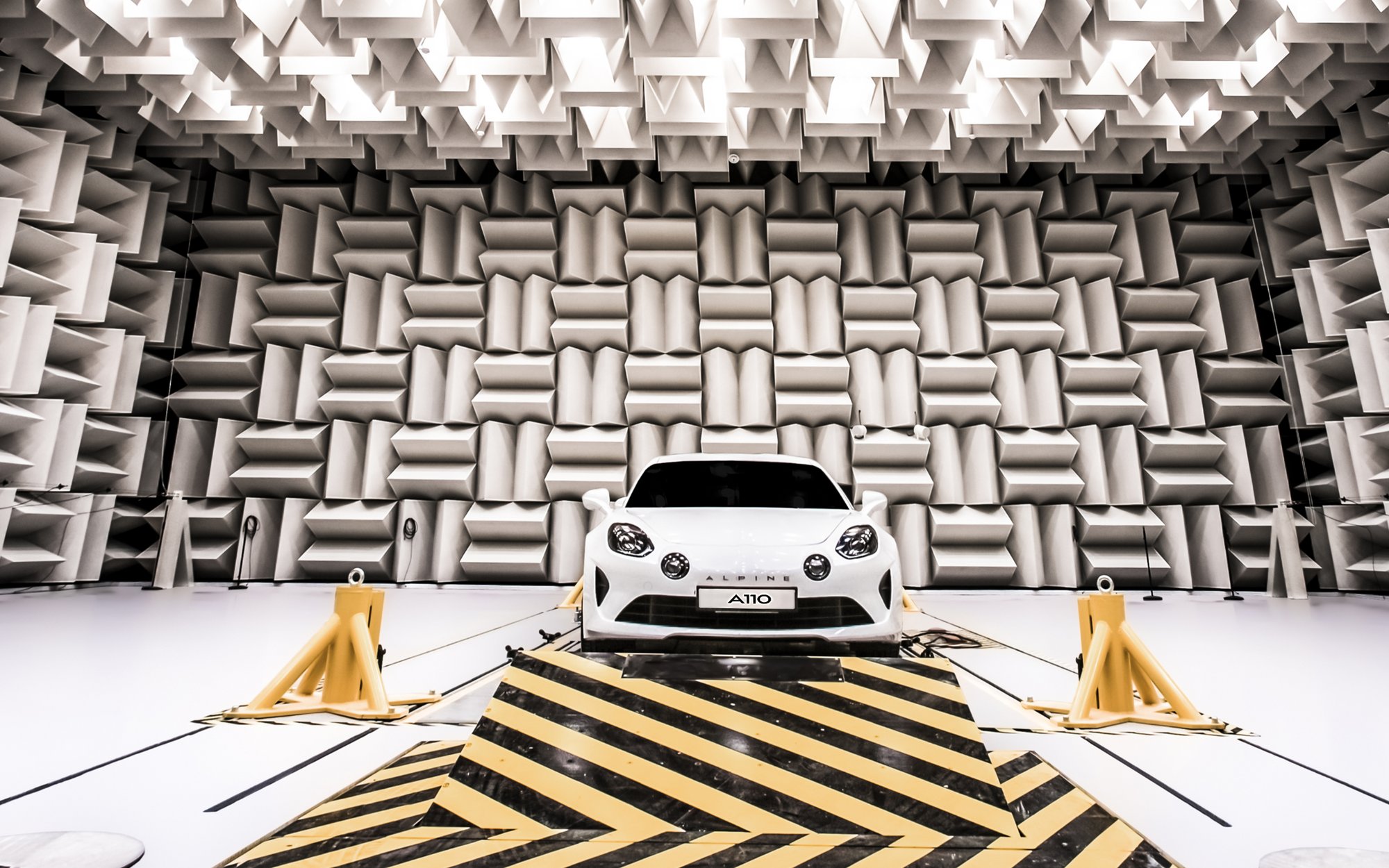 A white sports car coupe is positioned in a futuristic-looking room with sound-absorbing wall and ceiling panels. The vehicle rests on a yellow and black striped platform.