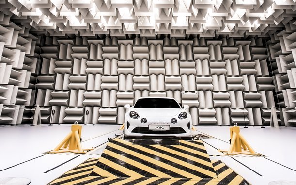 A white sports car coupe is positioned in a futuristic-looking room with sound-absorbing wall and ceiling panels. The vehicle rests on a yellow and black striped platform.