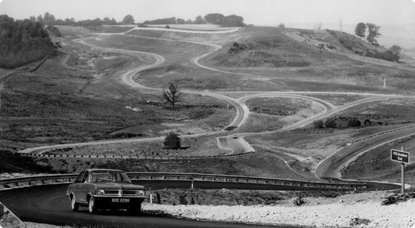 Une image historique en noir et blanc d'un circuit de course ou d'une piste d'essai vallonnée et sinueuse avec une voiture roulant au premier plan et d'autres pistes s'étendant à travers le paysage.