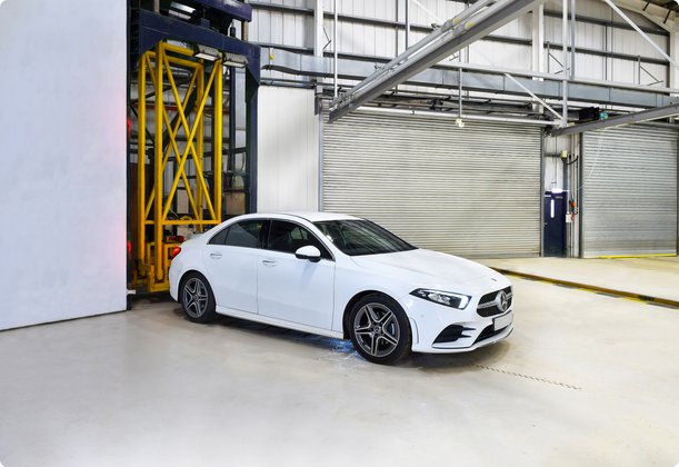 An elegant white coupe, likely a Mercedes-Benz, is positioned on a platform at the entrance of a wind tunnel or testing facility. The vehicle appears ready for aerodynamic testing.