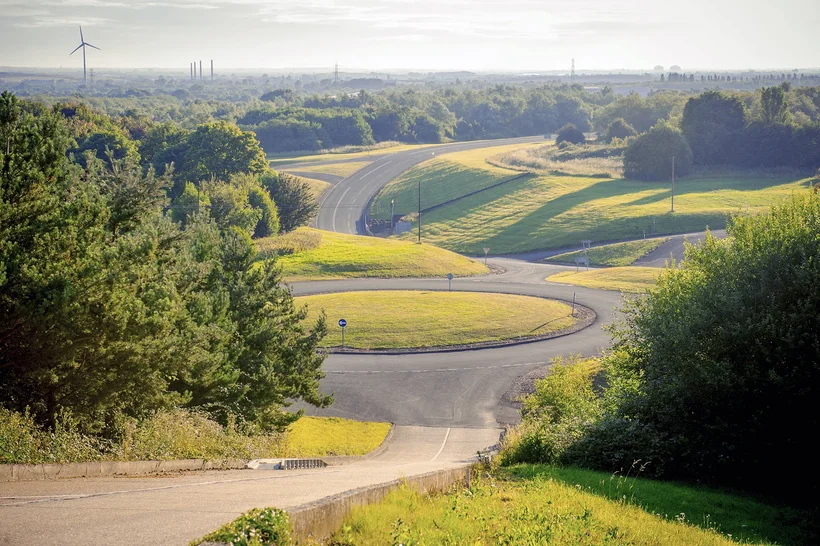 An elevated view of a test track or course that winds with multiple curves and inclines through a green, hilly landscape. The track is surrounded by trees and sun-drenched grassy areas. On the horizon, the outlines of a distant city or industrial complex are discernible.