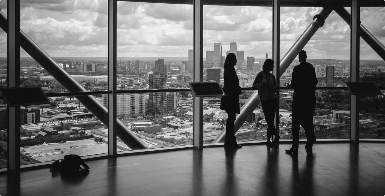 A black and white image showing the silhouettes of three people standing by large windows, looking out at an expansive city skyline with skyscrapers and a river. The windows have a diagonal grid structure.