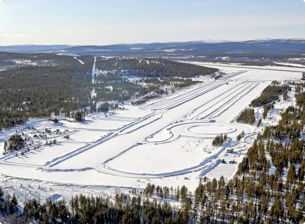 An aerial view of a vast snow-covered landscape, possibly a winter test track or a ski resort, with multiple parallel tracks cut into the snow, winding trails, and dense evergreen forests bordering the open areas.