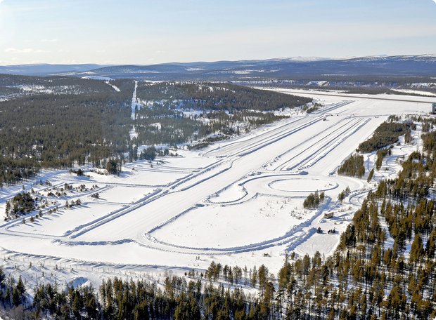 An aerial view of a vast snow-covered landscape, possibly a winter test track or a ski resort, with multiple parallel tracks cut into the snow, winding trails, and dense evergreen forests bordering the open areas.
