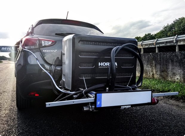 The rear view of a car with an attached, black emissions measurement device or test rig on the road, indicating real-world exhaust testing or driving trials. A sign reading "FUTAC CERAM" is also visible.