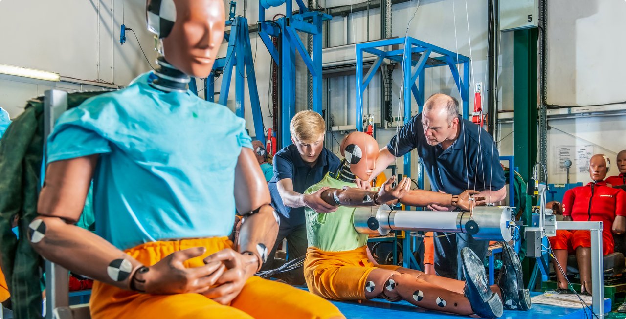 Close-up of a crash test dummy in a turquoise polo shirt and orange trousers, seated in a test rig with blue metal structures. Other dummies and technicians are visible in the background.