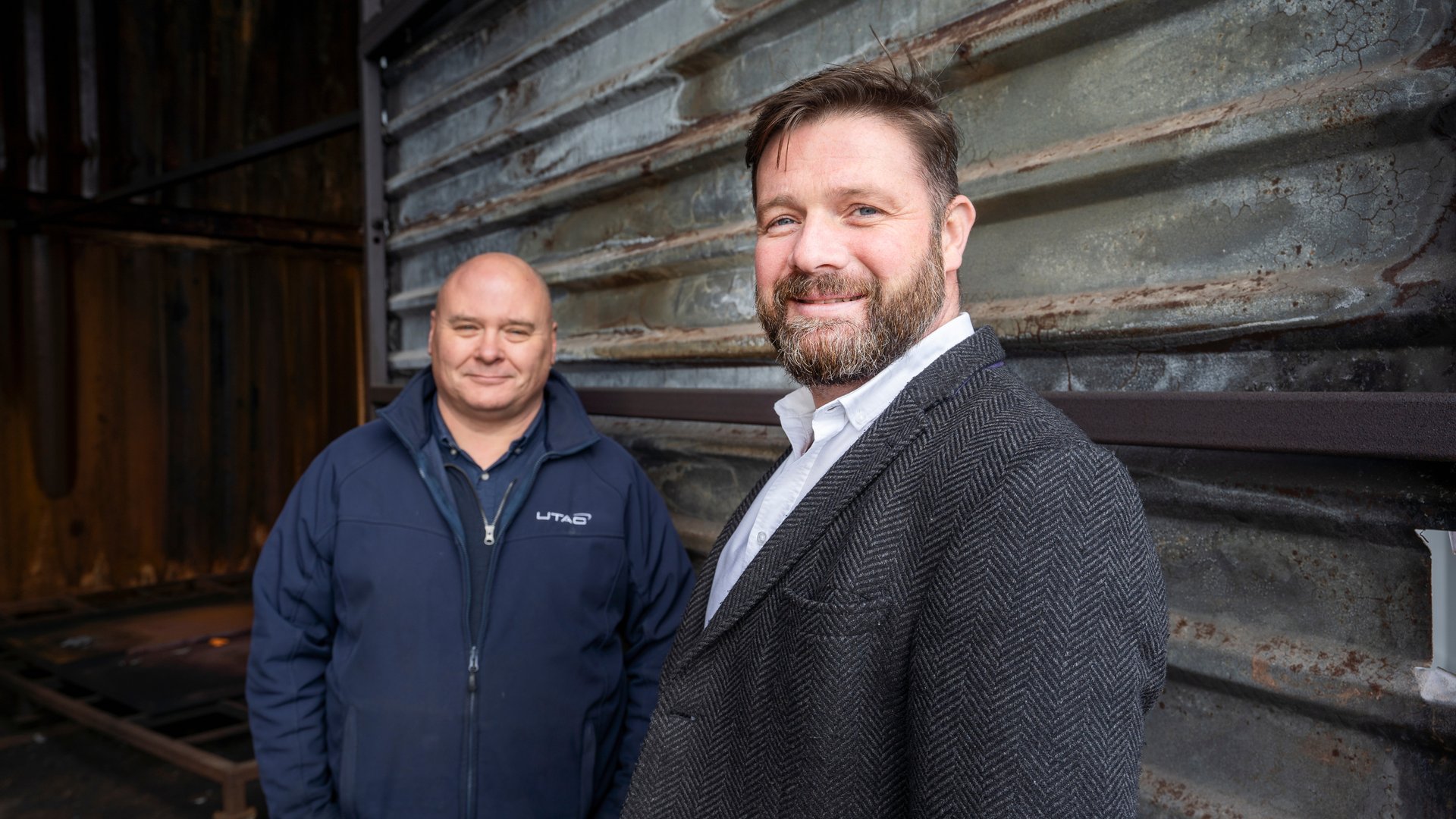 Two men in front of a metal wall – one in a suit, the other in workwear – representing leadership and craftsmanship.