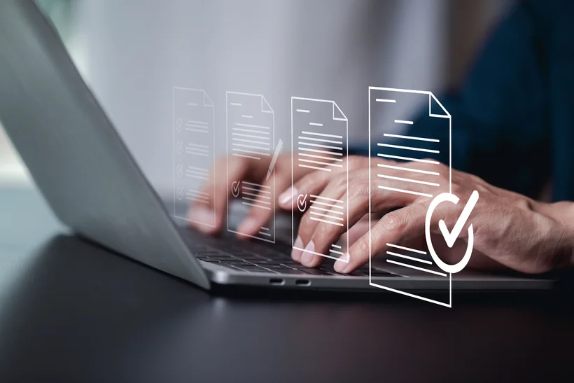 Close-up of a person's hands typing on a laptop keyboard. Above the keyboard and hands, holographic or digital representations of documents and a checkmark symbol are floating, indicating digital work, data processing, or document verification.